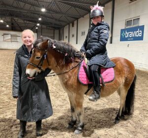 én ung jente står ved siden av en hest som har en annen jente på ryggen